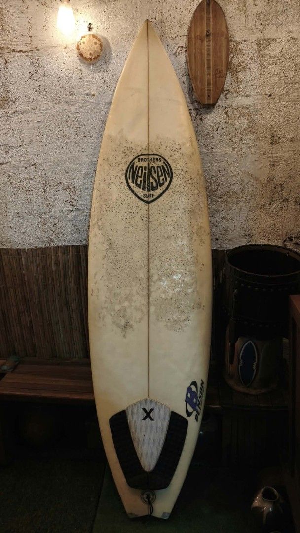 Brothers Neilsen Surf Surfboard (Category: Swim Trunks & Board Shorts) located in Manila. Submitted by glydelmalu86312 (Listing ID 1465320330, Image 1). Description: - Brand/Model: Brothers Neilsen surf board with Benson traction pad - Design: Classic white finish with central wooden stringer from nose to tail - Setup: Tri-fin configuration - Condition: Well-loved with wax residue, minor dings near the nose, a repaired patch near the logo, and a 