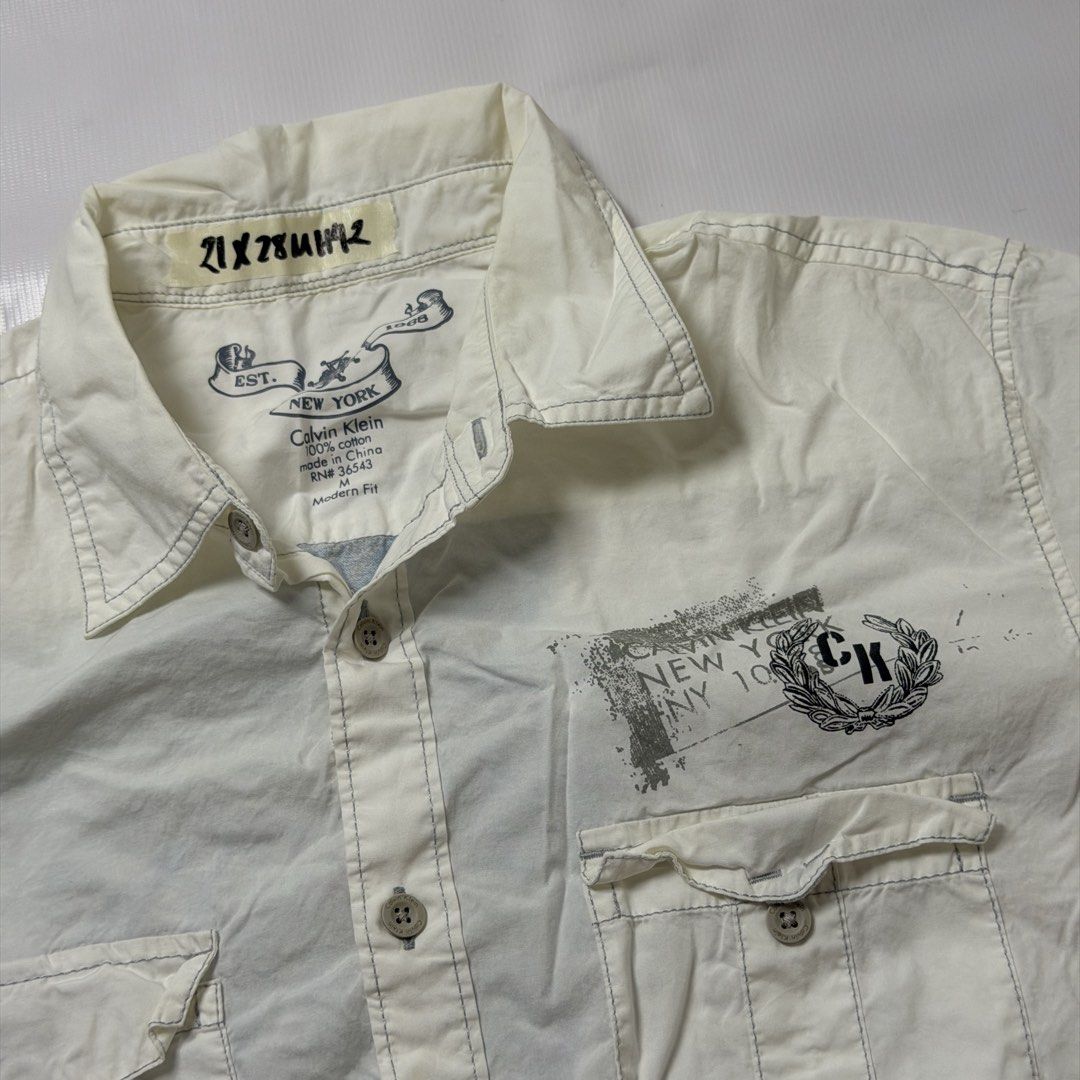 Calvin Klein Graphic Short Sleeve Button Down Shirt White Medium (Category: Tshirts & Polo Shirts ) located in Manila. Submitted by johnedel_james (Listing ID 1465280906, Image 2). Description: - Brand: Calvin Klein - Size: Medium (Modern Fit) - Material: 100% Cotton - Color: White - Details: Short sleeve, branded buttons, dual chest pockets with button flaps, small chest logo, and a bold graphic print on the back - Condition: Excellent, pre-love