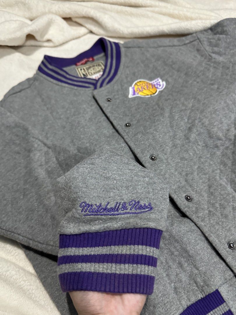 NBA Hardwood Classics by Mitchell & Ness (Category: Coats, Jackets and Outerwear) located in Albuera. Submitted by thriftanasias (Listing ID 1465331380, Image 3). Description: The image shows a gray Los Angeles Lakers snap-button varsity jacket featuring purple and gray striped ribbing on the collar, cuffs, and hem, along with the Lakers team logo patch on the chest..