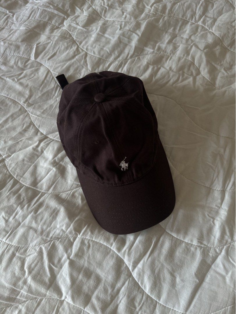 Polo Ralph Lauren Dark Brown Cap (Category: Caps & Hats) located in Naga. Submitted by maartsy.co (Listing ID 1465272014, Image 1). Description: - Brand: Polo Ralph Lauren - Color: Dark Brown with white embroidered logo - Design: Classic 6-panel construction with a curved brim - Fit: Adjustable strap at the back for a custom fit - Condition: Clean fabric, minimal wear, well-maintained.