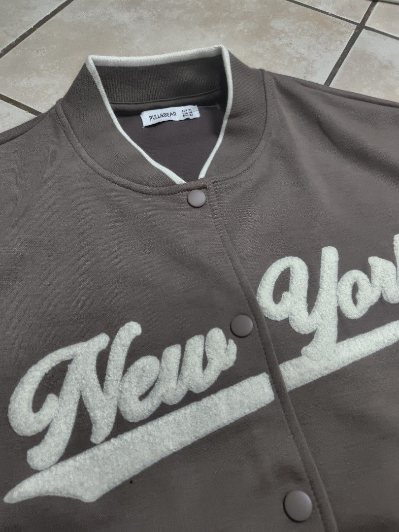 Pull&Bear New York Varsity Jacket Brown (Category: Coats, Jackets and Outerwear) located in Manila. Submitted by safetiger.42d40e (Listing ID 1465323737, Image 2). Description: - Brand: Pull&Bear - Color: Brown with cream and white details - Style: Varsity jacket with snap button closure - Design: 'New York' chenille embroidered lettering on the chest - Details: Ribbed collar, cuffs, and hem with white tipping; two front pockets - Fit & Material: