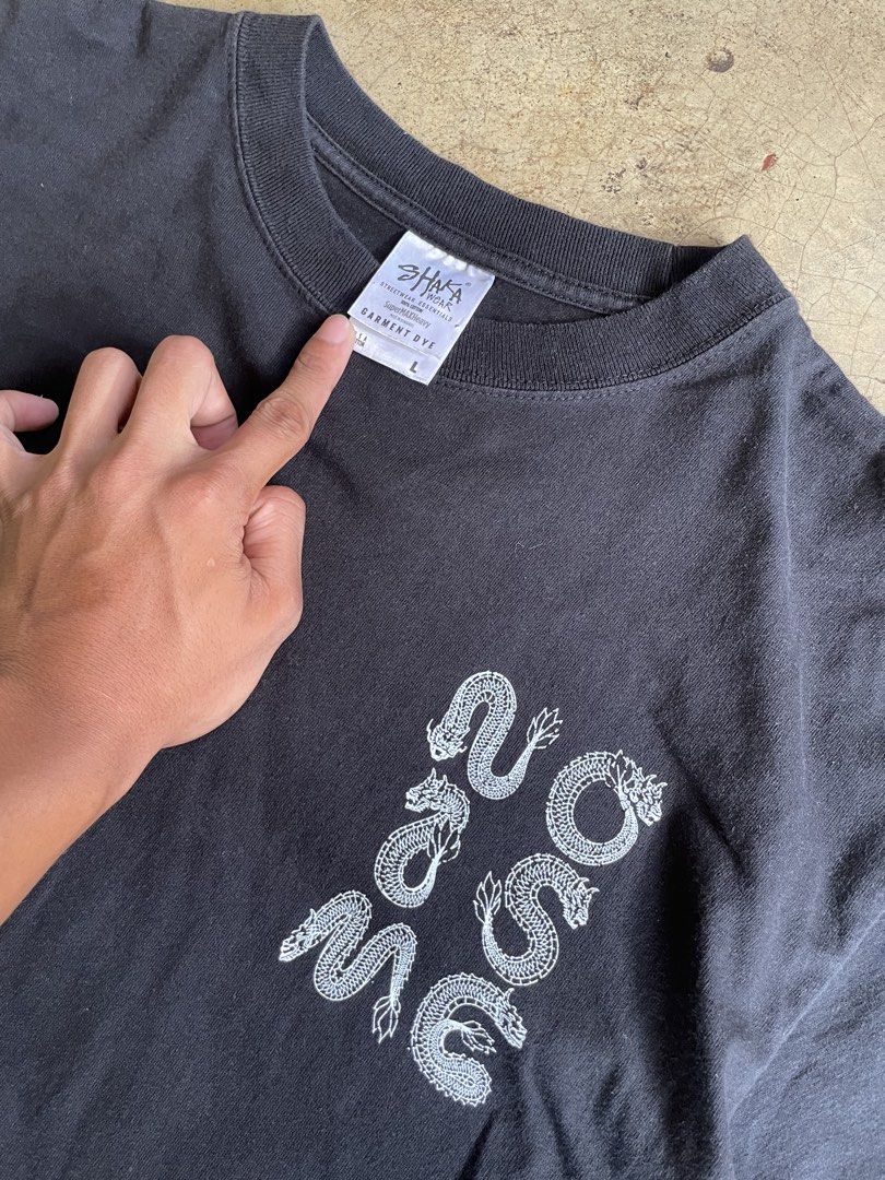 SHAKA WEAR (L) (Category: Activewear) located in Valencia. Submitted by macsss11 (Listing ID 1465192558, Image 3). Description: The image shows a black pigment-dyed t-shirt featuring a white graphic print on the front and a Shaka Wear neck label..