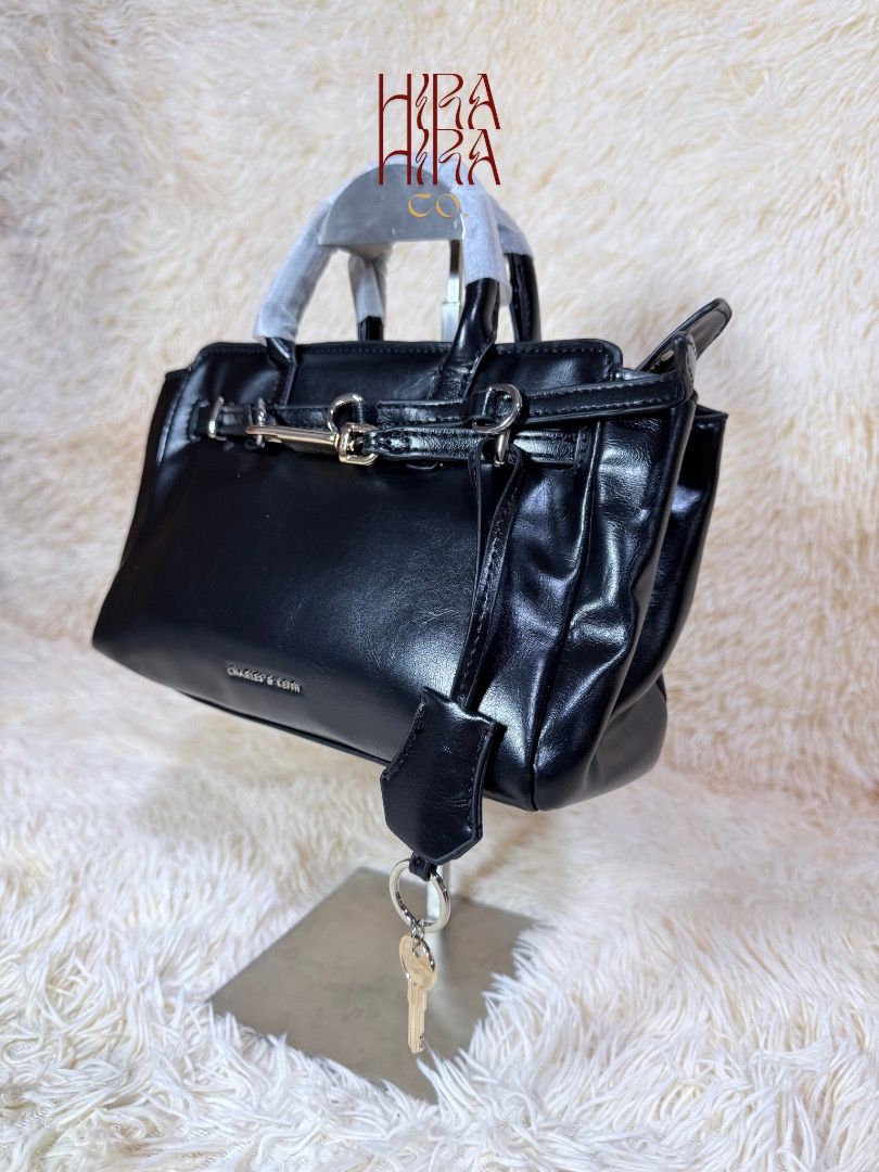 Charles & Keith Black Handbag (Category: Luxury Bags & Wallets) located in Caloocan City. Submitted by hirahira.co (Listing ID 1467082592, Image 2). Description: - Brand: Charles & Keith - Color & Finish: Black with a smooth, glossy finish - Hardware: Silver-tone hardware - Handles: Dual top handles (still wrapped in protective foam) - Details: Decorative front belt detail and attached padlock charm with keys - Compartment: Secure zippered main c