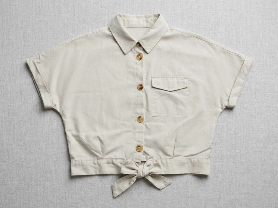 Cream button-down crop top with tie front (Category: Babies & Kids Fashion) located in Manila. Submitted by bright.atina14 (Listing ID 1467073027, Image 1). Description: - Color: Cream - Style: Short-sleeve button-down with collared neckline - Details: Tortoiseshell-style buttons, single left chest pocket, cuffed sleeves, and a trendy tied knot at the hem - Material: Lightweight and comfortable fabric - Styling Tip: Pairs perfectly with denim sho