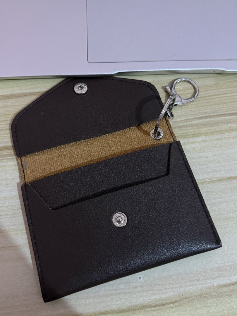Dark brown leather card holder pouch with key ring (Category: Wallets & Card holders) located in Manila. Submitted by swifthorse.a59ed4 (Listing ID 1467015440, Image 2). Description: - Color/Material: Dark brown faux leather - Design: Envelope style flap with snap button closure - Branding: Debossed "de." logo on the front - Hardware: Attached silver keyring and clasp inside - Interior: Brown fabric lining across the middle - Use: Compact size pe