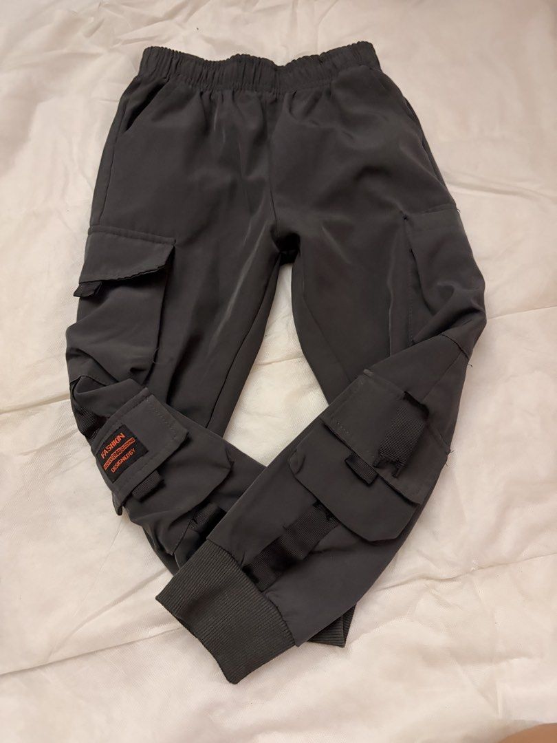 Dark Grey Cargo Pants with Multiple Pockets and Cuffs (Category: Babies & Kids Fashion) located in Dasmarinas. Submitted by thriftcavite (Listing ID 1466987220, Image 1). Description: - Color: Dark grey - Style: Streetwear cargo pants - Pockets: Multiple utility pockets with straps and buckle accents - Details: Small orange text patch on the lower left pocket - Fit: Elastic waistband and ribbed ankle cuffs for maximum comfort.