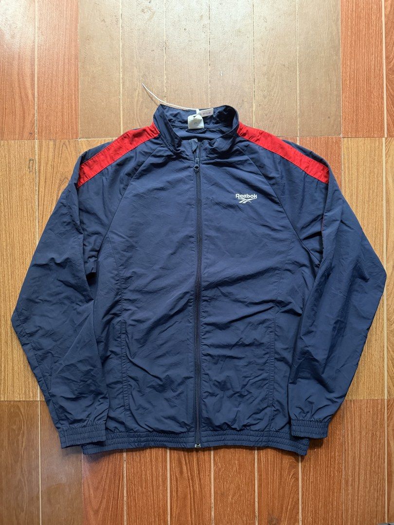 Reebok Navy Blue and Red Track Jacket (Category: Coats, Jackets and Outerwear) located in Dasmarinas. Submitted by kylebecker (Listing ID 1467001120, Image 1). Description: - Color: Navy blue with red panels across the shoulders - Branding: Embroidered white Reebok logo on the right chest - Closure & Fit: Full zip front with elastic cuffs and hem - Material: Lightweight fabric - Condition: Clean stitching throughout, perfect for workouts or casua