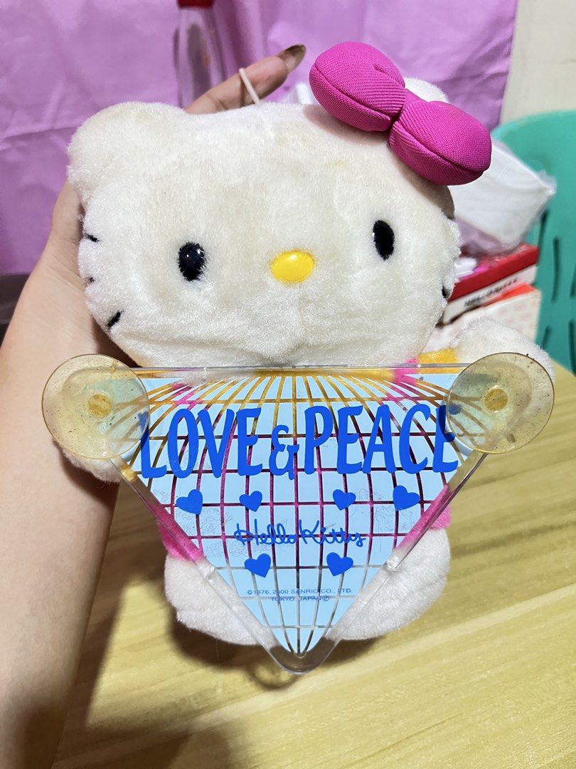 Sanrio Hello Kitty Love & Peace Plush Toy (Category: Plushies & Soft Toys) located in Manila. Submitted by janetecson05 (Listing ID 1467002721, Image 1). Description: - Official Sanrio Kitty plush toy (Dated 1976, 2000) - Features classic white Kitty with a bright pink bow - Holding a triangular acrylic sign with 'LOVE & PEACE' design, blue grid, and hearts - Includes 2 suction cups on the sign corners for easy hanging on car windows or glass - P