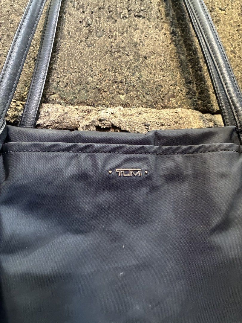 Tumi Black Nylon Tote Bag (Category: Tote Bags) located in Manila. Submitted by assortedgoodsv3 (Listing ID 1467165809, Image 2). Description: - Brand: Tumi - Material: Durable black nylon with leather straps and trim - Closure: Zippered top for security - Exterior: Front pocket with metal Tumi logo plaque and hanging leather luggage tag with gold hardware - Hardware: YKK zipper pull on the interior - Condition: Shows normal wear on hardware plat