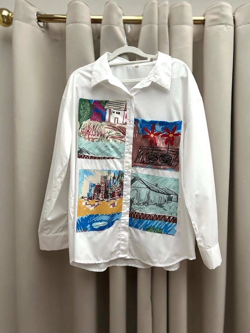 White Button-Up Shirt With Artistic Patchwork Prints (Category: Shirts) located in Cainta. Submitted by unboxingstuff (Listing ID 1467020266, Image 1). Description: - Color: Crisp white with vibrant, multi-colored illustrated patch pockets - Design: Features bright landscape and cityscape drawings in blue, red, green, and yellow - Details: Classic collar, long sleeves, and front button-down closure - Material: Smooth, lightweight, and comfortable