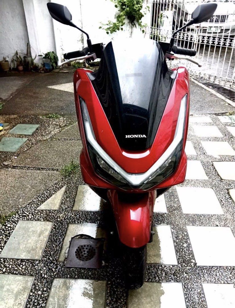 2025 Honda PCX 160 (Category: Motorbikes for Sale) located in Antipolo. Submitted by goldenmoon_33cc19 (Listing ID 1467177500, Image 1). Description: Red Honda PCX 160 scooter, black windshield and mirrors. Clean body panels, dark alloy wheels. Dual headlights with LED strips. Comfortable black seat, standard exhaust pipe. Street-ready commuter bike parked outdoors..