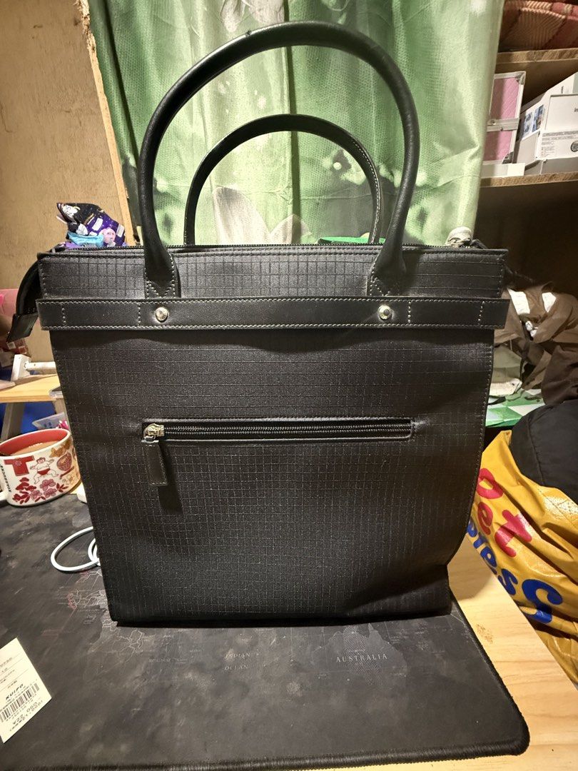 Anne Klein Black Tote Bag (Category: Tote Bags) located in Opol. Submitted by great_finds_kollexion (Listing ID 1467171733, Image 2). Description: Black structured tote bag by Anne Klein. Grid texture pattern on fabric exterior. Double handles with some wear along the edges visible in photos. Horizontal decorative strap across the front featuring the brand name embossed on metal-accented hardware. Exterior back pocket with zipper closure. Fabric 