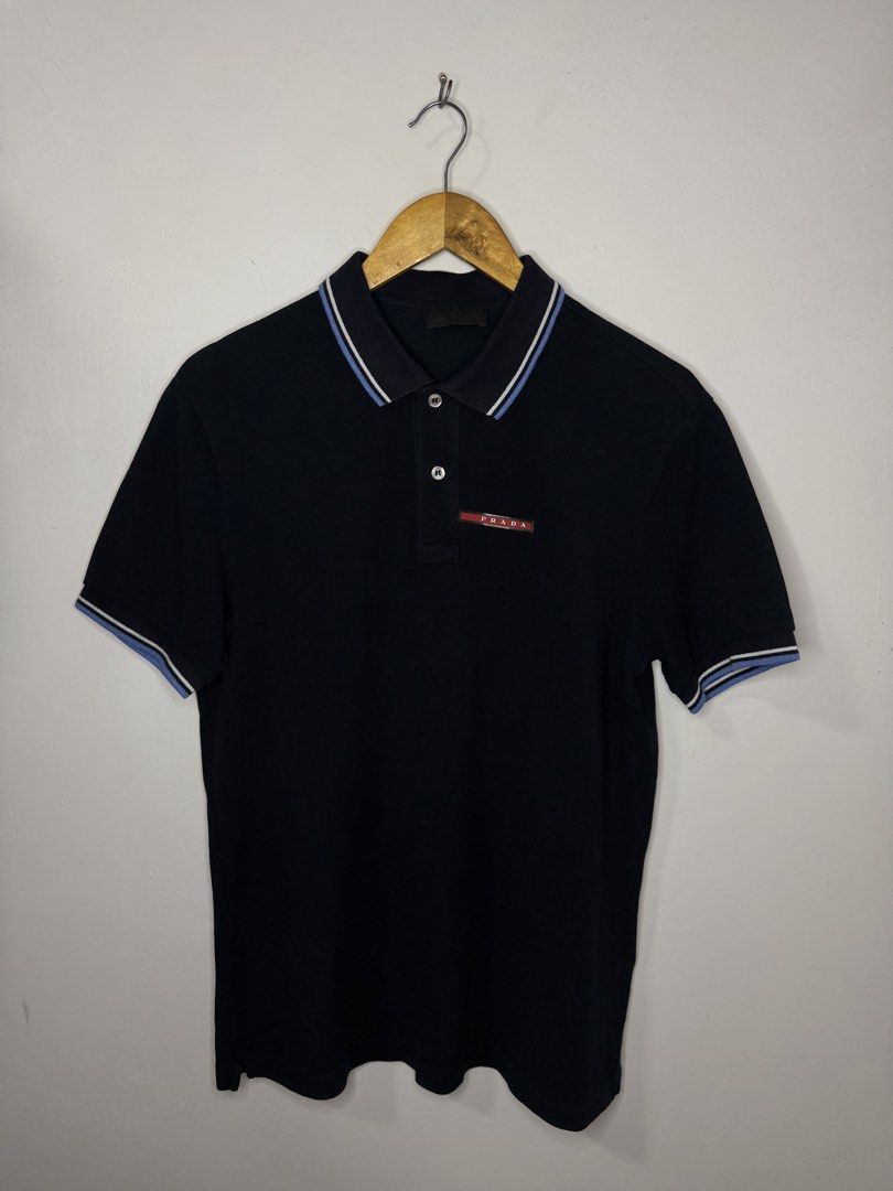 Prada Polo Shirt (Dark Blue) (Category: Tshirts & Polo Shirts ) located in Manila. Submitted by jcarchives14 (Listing ID 1467278168, Image 1). Description: - Brand: Prada - Color: Solid black body with contrasting blue and white striped collar and cuffs - Style: Short sleeve polo shirt with a classic two-button placket - Details: Premium mother-of-pearl buttons and iconic red Prada logo label on the left chest - Material/Finish: Ribbed knit colla