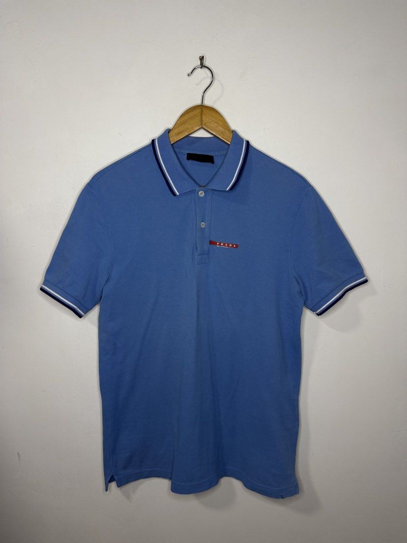 Prada Polo Shirt (Light Blue) (Category: Tshirts & Polo Shirts ) located in Manila. Submitted by jcarchives14 (Listing ID 1467277848, Image 1). Description: - Authentic Prada polo shirt in light blue - Premium soft cotton fabric construction - Contrast navy blue and white striping on collar and sleeve cuffs - Classic two-button placket closure - Signature red rectangular Prada logo patch on the left chest - Clean solid back and body design - Perf