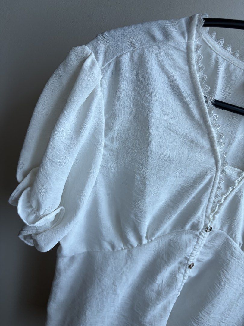 Redgirl White Blouse Size L (Category: Blouses) located in Makati City. Submitted by biancabuysells (Listing ID 1467245658, Image 3). Description: - Brand: Redgirl - Size: Large (L) but fits medium - Color: Classic White - Design: V-neck with delicate lace trim & metallic front buttons - Sleeves: Short puff sleeves with ruffled cuffs - Fit: Fitted underbust with a relaxed lower hem - Fabric: Lightweight textured material with a subtle pattern - S