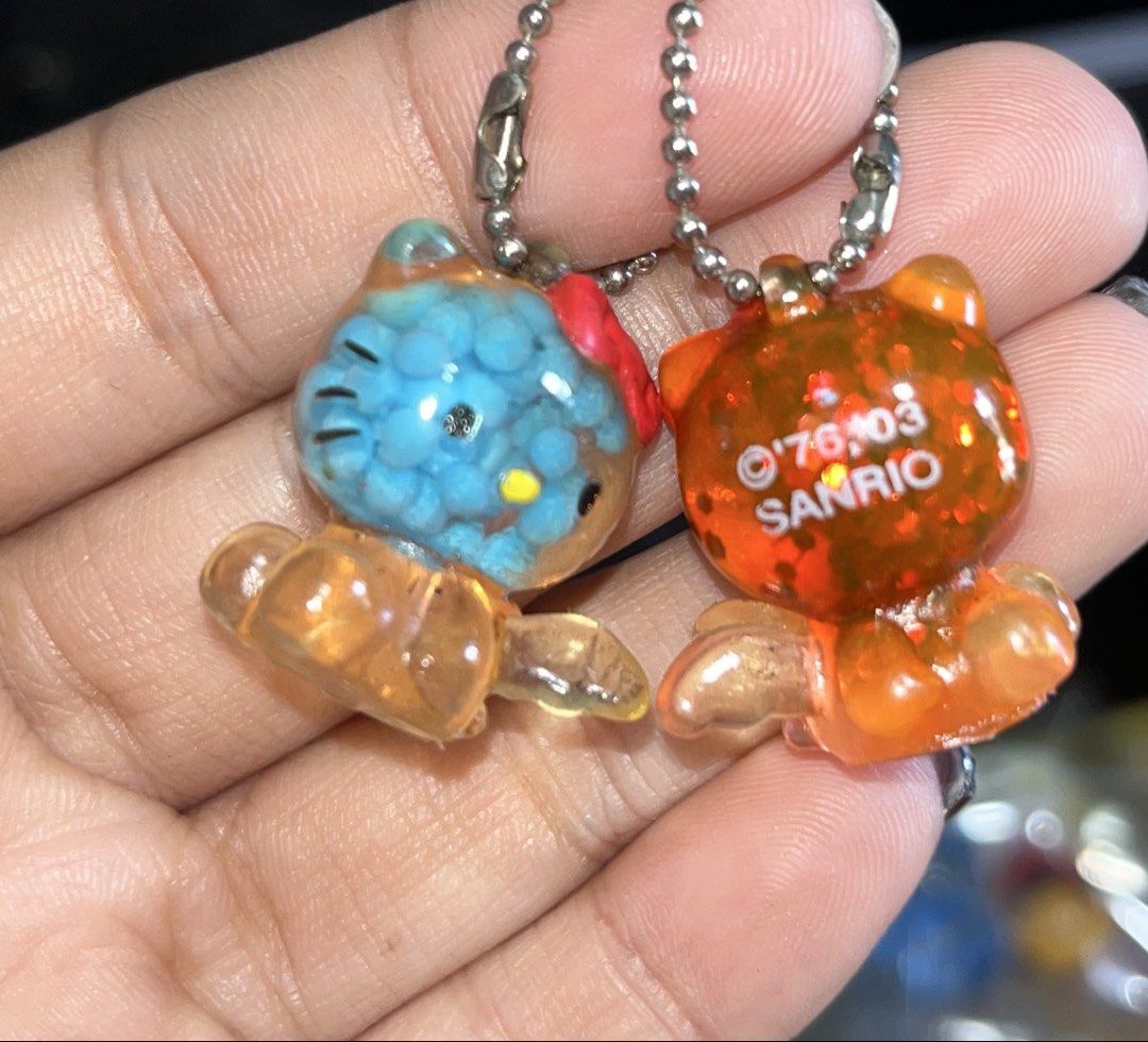 Sanrio Hello Kitty Resin Bead and Glitter Keychains Pair (Category: Character Goods) located in Manila. Submitted by janetecson05 (Listing ID 1467309356, Image 2). Description: - Set of 2 authentic Sanrio Kitty keychains - Features silver ball chains for easy hanging - Blue keychain: Clear resin filled with floating small round beads - Orange keychain: Glittery figure with red bow and angel wings - Both feature suspension liquid inside clear body