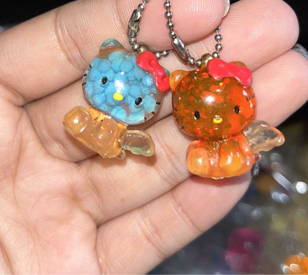 Sanrio Hello Kitty Resin Bead and Glitter Keychains Pair (Category: Character Goods) located in Manila. Submitted by janetecson05 (Listing ID 1467309356, Image 1). Description: - Set of 2 authentic Sanrio Kitty keychains - Features silver ball chains for easy hanging - Blue keychain: Clear resin filled with floating small round beads - Orange keychain: Glittery figure with red bow and angel wings - Both feature suspension liquid inside clear body