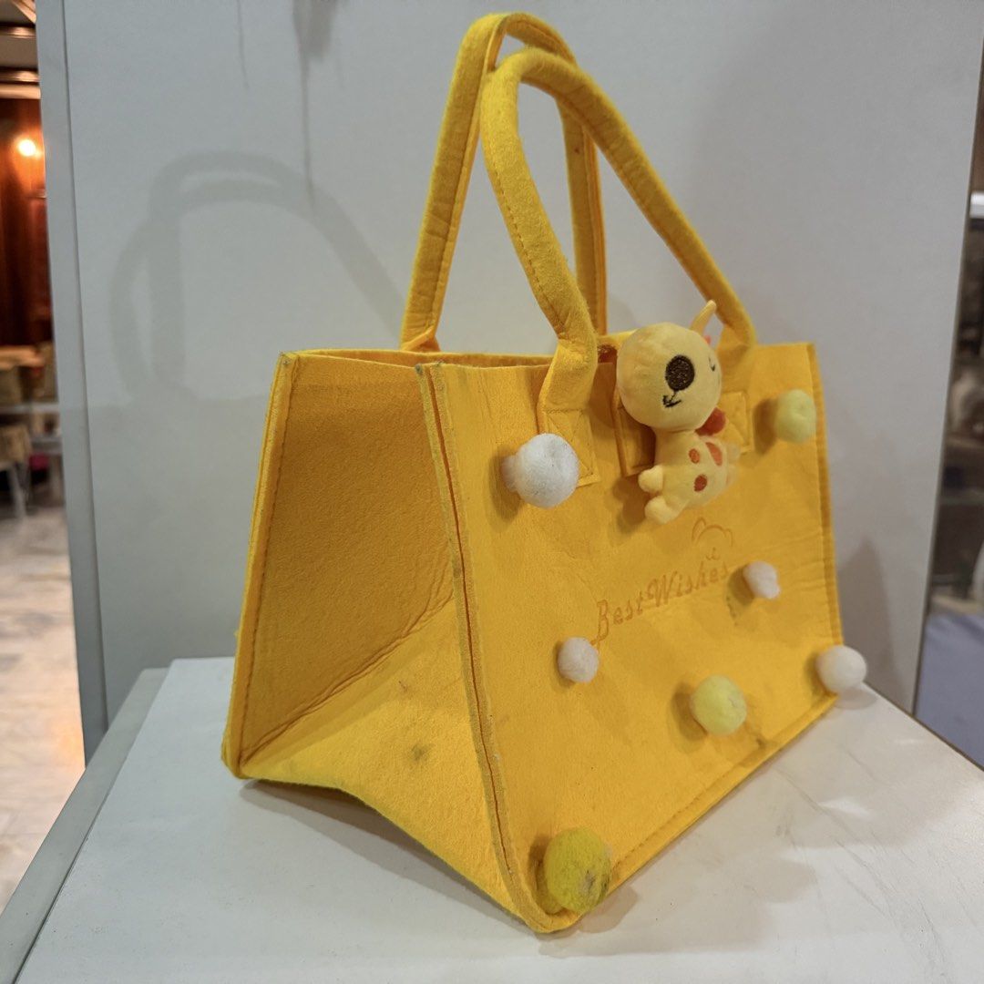 Yellow Felt Tote Bag with Cute Giraffe Design (Category: Tote Bags) located in Marikina City. Submitted by lovegoeson (Listing ID 1467289239, Image 2). Description: - Color/Material: Bright yellow felt tote bag - Cute Details: Features an attached stuffed giraffe, white and yellow pom-pom balls, and embroidered "Best Wishes" text - Design: Twin top handles with a convenient open top - Condition: Some light dirt marks on the exterior body and bott