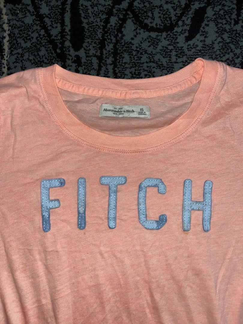 Abercrombie & Fitch “Fitch” Shirt (Category: Shirts) located in Manila. Submitted by kashjulliane (Listing ID 1467366611, Image 2). Description: - Brand: Abercrombie & Fitch - Size: XS - Color: Pink with light blue 'FITCH' appliques - Material: Soft slub knit fabric - Design: Short sleeves, round neckline - Origin: Made in Cambodia - Condition: Excellent, casual everyday wear width 16 length 25.