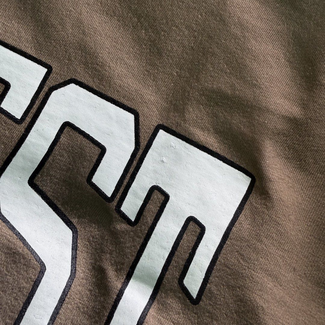 Coziest Brown Graphic Tee Size M (Category: Shirts) located in Benito Soliven. Submitted by markedasdifferent (Listing ID 1467521102, Image 2). Description: - Brand: Coziest - Size: Medium - Color: Brown with bold white collegiate chest graphic - Material: 80% cotton, 20% polyester (pre-shrunk) - Care: Machine wash cold - Condition: Clean, neat stitching, with woven hem label.
