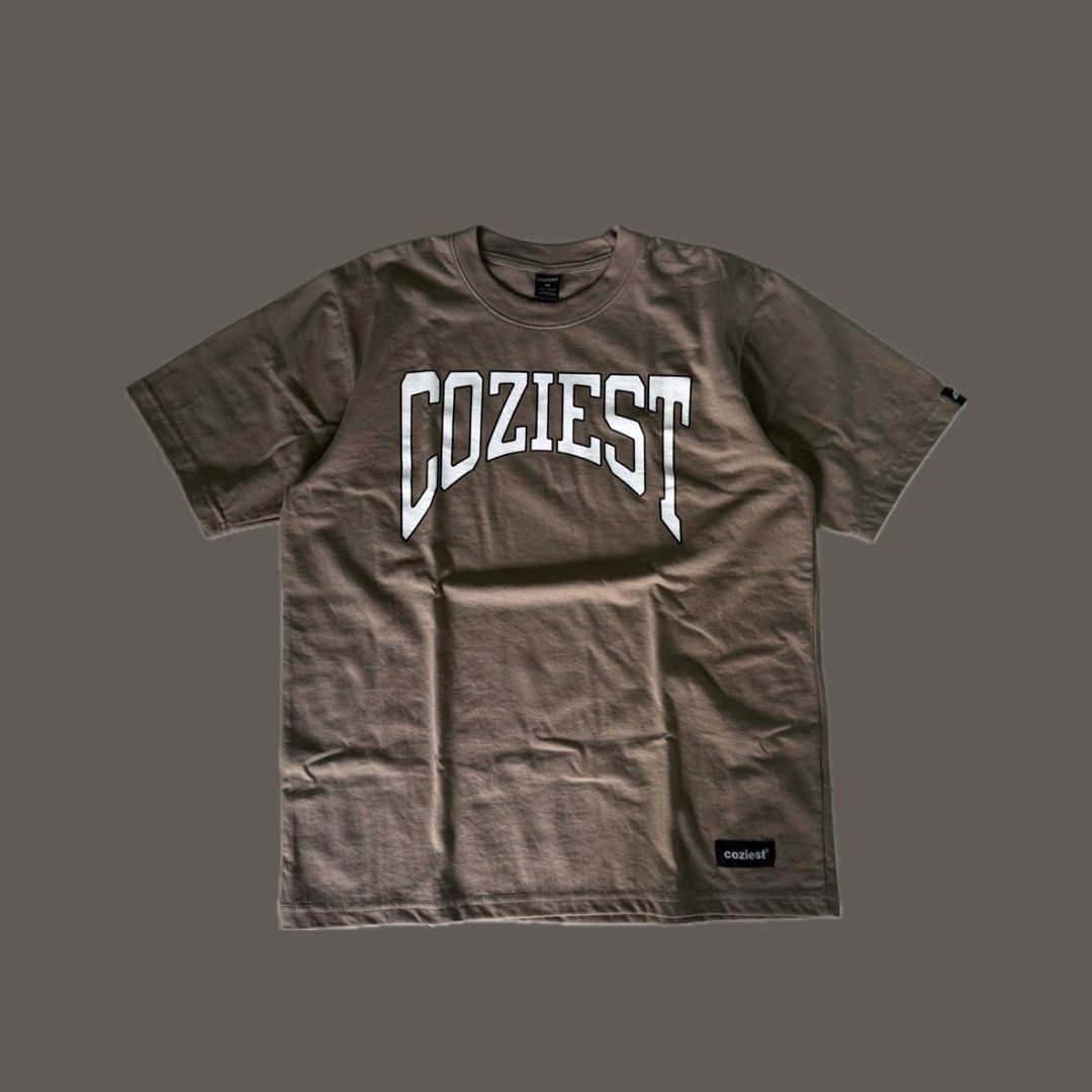 Coziest Brown Graphic Tee Size M (Category: Shirts) located in Benito Soliven. Submitted by markedasdifferent (Listing ID 1467521102, Image 1). Description: - Brand: Coziest - Size: Medium - Color: Brown with bold white collegiate chest graphic - Material: 80% cotton, 20% polyester (pre-shrunk) - Care: Machine wash cold - Condition: Clean, neat stitching, with woven hem label.