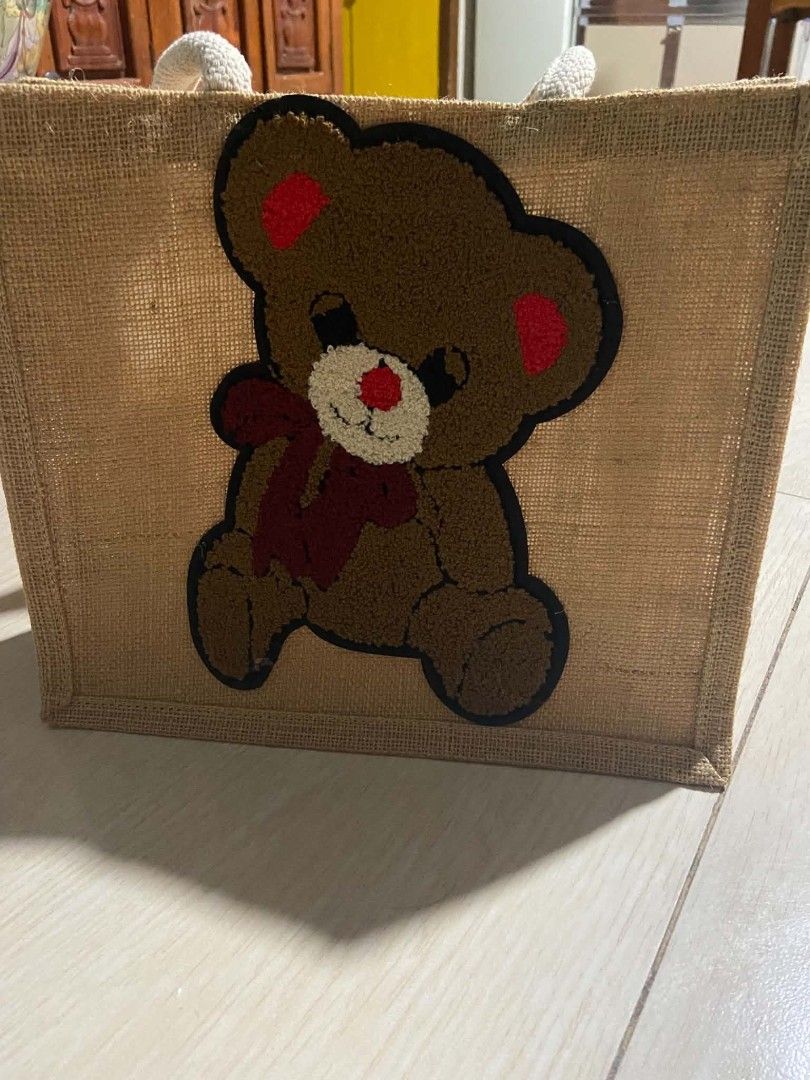 Cute Teddy Bear Jute Tote Bag (Category: Tote Bags) located in Zaragoza. Submitted by sherree1281 (Listing ID 1467517015, Image 1). Description: - Material: Natural tan burlap fabric body - Design: Large fuzzy teddy bear patch with red inner ears, red nose, and a maroon bow - Handles: Thick and sturdy rope handles at the top - Structure: Structured square shape for easy storage - Condition: Brand new - Perfect for daily errands, casual outings, o