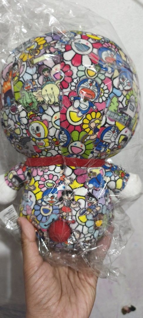 Doraemon Takashi Murakami Collab Plush Toy (Category: Plushies & Soft Toys) located in Manila. Submitted by jelaibinzz (Listing ID 1467367821, Image 2). Description: - Doraemon plush toy featuring iconic Takashi Murakami colorful floral and character prints - Classic details: red collar with yellow bell, red nose, and round red tail - Condition: Brand new, still sealed inside clear plastic wrapper - Must-have collector's piece for Doraemon fans a
