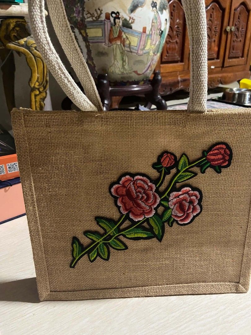 Jute Tote Bag with Embroidered Floral Patch (Category: Tote Bags) located in Zaragoza. Submitted by sherree1281 (Listing ID 1467517131, Image 1). Description: - Material: Natural brown burlap with thick woven canvas handles - Design: Large embroidered red rose patch with green stem and leaves on the front - Structure: Rectangular shape with reinforced borders for durability - Capacity: Roomy interior, perfect for daily essentials or grocery runs 
