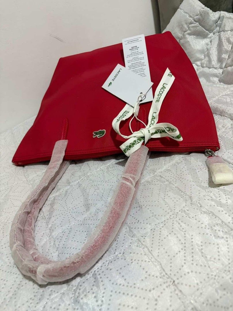 Lacoste Red Tote Bag With Ribbon Accent (Category: Tote Bags) located in Polomolok. Submitted by legadad94559 (Listing ID 1467566016, Image 3). Description: - Authentic Lacoste tote bag in a vibrant red canvas - Brand new with original retail tags attached - Secure top zipper closure - Sturdy shoulder straps still wrapped in protective plastic - Elegant branded ribbon tied neatly on the front handle - Spacious interior featuring a zippered pocket