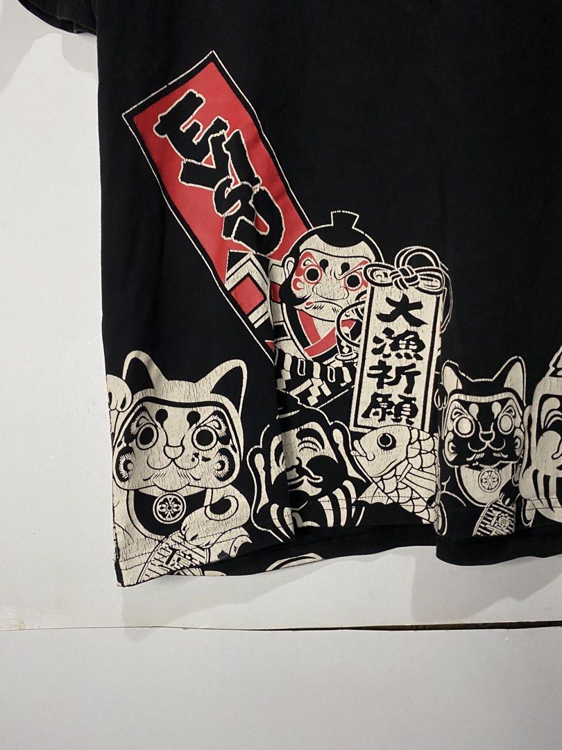 TSHIRT (Category: Tshirts & Polo Shirts ) located in Jaen. Submitted by oshimamark58418 (Listing ID 1467516398, Image 3). Description: - Brand: EVISU - Color: Black with white and red Japanese graphic prints - Size: L (Tag says XL, measured 21x28 inches) - Design: Daruma dolls, lucky cats, and Japanese text graphics along the hem, with a red chest logo and branded back collar - Condition: 7/10 pre-loved condition - Style: Casual streetwear.