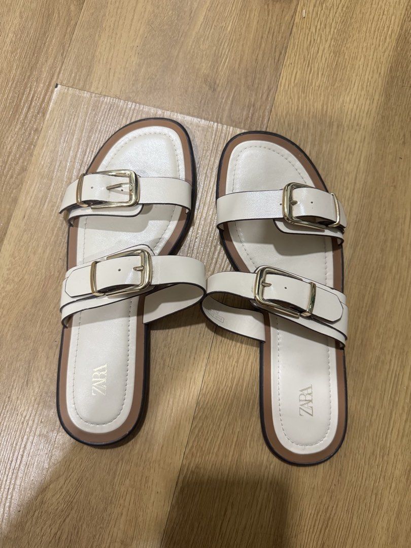Zara White Double Buckle Flat Sandals Size 37 (Category: Flats & Sandals) located in Mandaluyong City. Submitted by piadeclutters (Listing ID 1467587638, Image 1). Description: White flat sandals from Zara. Double strap design with gold-tone metal buckles. Cream-colored footbed with brown trim. Size 37 EUR. Textured black rubber soles underneath showing minimal wear. Very clean overall condition. - used less than 5 times only in malls - comfy fit