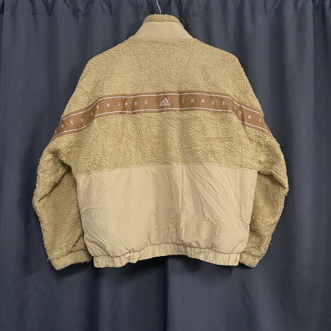 Adidas Beige Sherpa Fleece Jacket (Category: Coats, Jackets and Outerwear) located in Santa Maria. Submitted by saintarchive316 (Listing ID 1467835697, Image 3). Description: - Brand: Adidas - Material: Fluffy sherpa fleece with smooth nylon lower panels - Color: Beige with distinctive brown stripe - Details: High neck collar, zipper with snap-button closure, and side pockets - Branding: 'THREE STRIPES' lettering and logo across the back - Condit