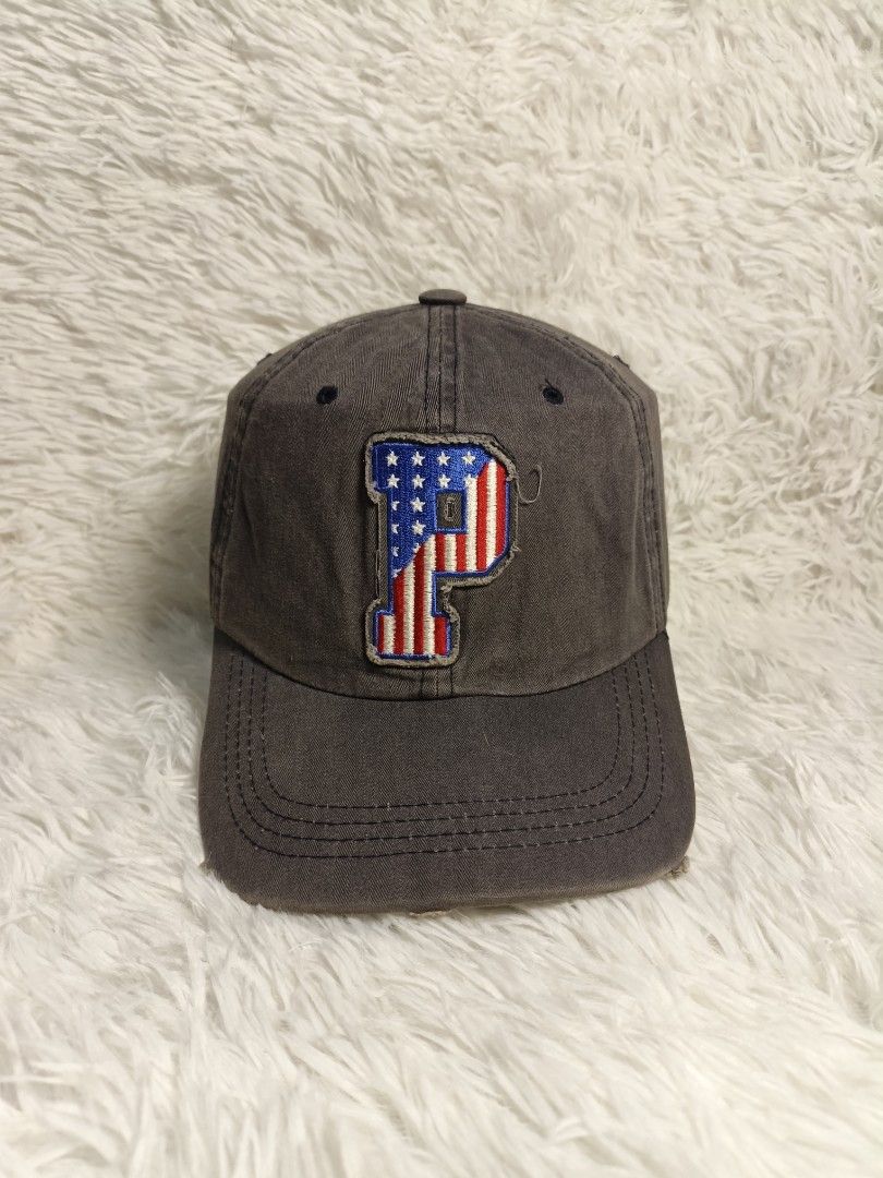 Authentic Pony Dark Grey Baseball Cap With American Flag P Patch (Category: Caps & Hats) located in Imus. Submitted by grindhouse.thriftshop (Listing ID 1467737384, Image 1). Description: - Color: Dark grey with a trendy distressed look - Design: Embroidered 'P' patch with USA flag fill on the front - Build: Classic 6-panel structure with top button and breathable eyelets - Brim: Curved brim with stylish intentional fraying along the edge - Style