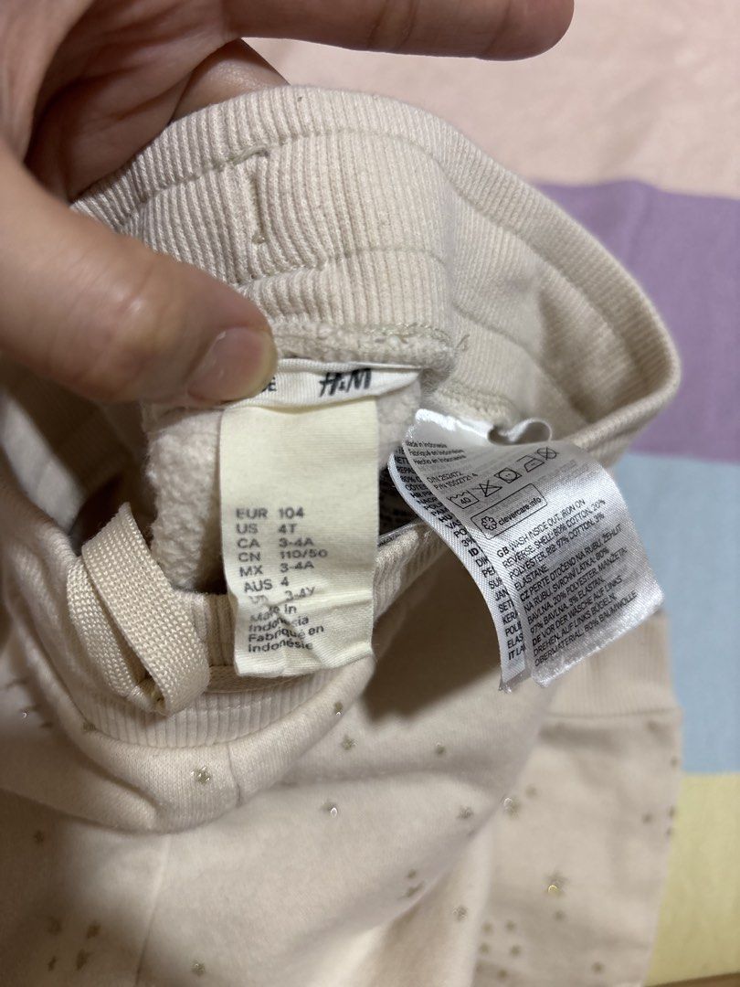 H&M Star Print Sweatpants Joggers (Category: Babies & Kids Fashion) located in Manila. Submitted by wardrobeinmnl (Listing ID 1467690981, Image 3). Description: - Brand/Item: H&M Beige Sweatpants with Sparkly Gold Stars - Size: EUR 104, US 4T, 3-4 Years - Material: Comfortable cotton blend with soft fleece lining - Features: Elastic waistband, adjustable drawstring, and ribbed ankle cuffs - Condition: Pre-loved with a slight pink stain near the f