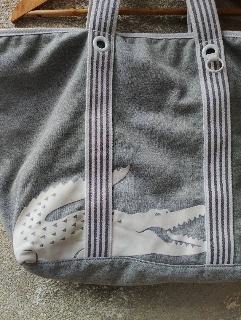 Lacoste Grey Tote Bag with Pouch (Category: Tote Bags) located in Iligan City. Submitted by norhazimmu53721 (Listing ID 1467625865, Image 2). Description: - Color: Classic Grey with bold striped shoulder handles - Iconic Design: Oversized white Lacoste crocodile graphic on the front - Extra Value: Includes a matching small zippered pouch attached by a strap - Interior Features: Patterned green crocodile lining, one zip pocket, and a secure snap b