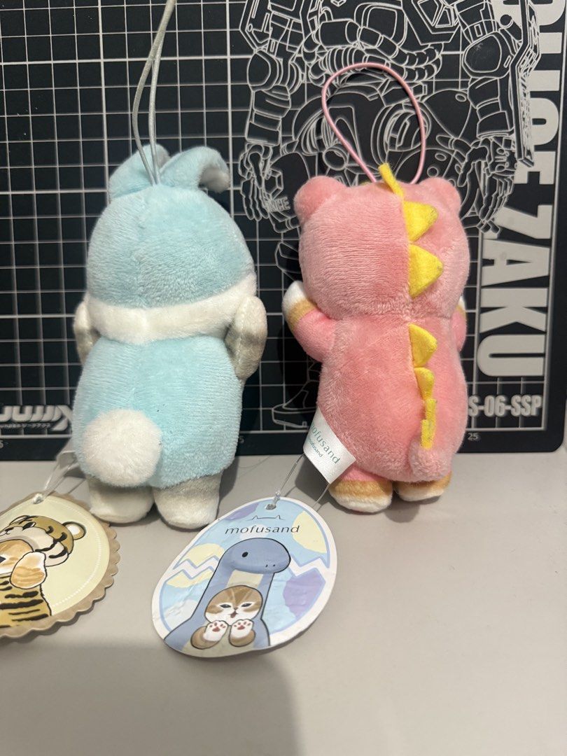 Mofusand Cat Plush Toy Keychain Set (Category: Plushies & Soft Toys) located in Victoria. Submitted by pisangpaulotoys (Listing ID 1467807059, Image 2). Description: - Set of 2 Mofusand cat plush keychains - Designs: Blue bunny suit and pink dinosaur suit - Made from ultra-soft, huggable fabric with high-quality embroidered facial features - Includes sturdy hanging loops for easy attachment to bags or keys - Brand new with original cardboard hang