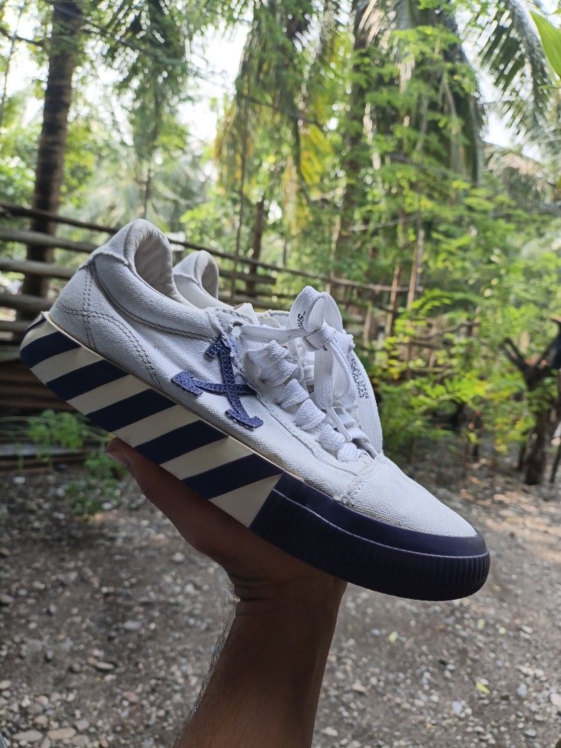Off-White Vulcanized Low Sneakers White Blue (Category: Sneakers) located in Maasim. Submitted by ukayganda25 (Listing ID 1467656829, Image 2). Description: - Brand: Off-White - Style: Low top sneakers in white canvas with iconic blue arrow details - Design: Vulcanized sole with text on bottom, 'SHOELACES' lettering on laces, and a purple hand graphic on the insoles - Condition: Pre-owned with some visible wear on the canvas and soles - Vibe: Per