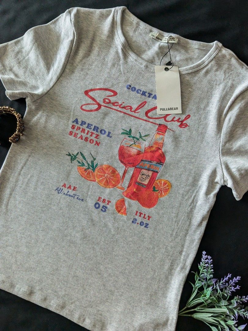 Pull & Bear Aperol Spritz Graphic Ribbed T-Shirt (Category: Shirts) located in Kananga. Submitted by aurylien.clo (Listing ID 1467612513, Image 1). Description: - Brand: Pull & Bear - Condition: Brand new with original store tag attached - Color: Light grey - Material: Comfortable ribbed cotton - Design: Cute Aperol Spritz graphic with bottle, cocktail glass, and orange slices - Details: Short sleeves, scoop neckline, red and blue lettering.