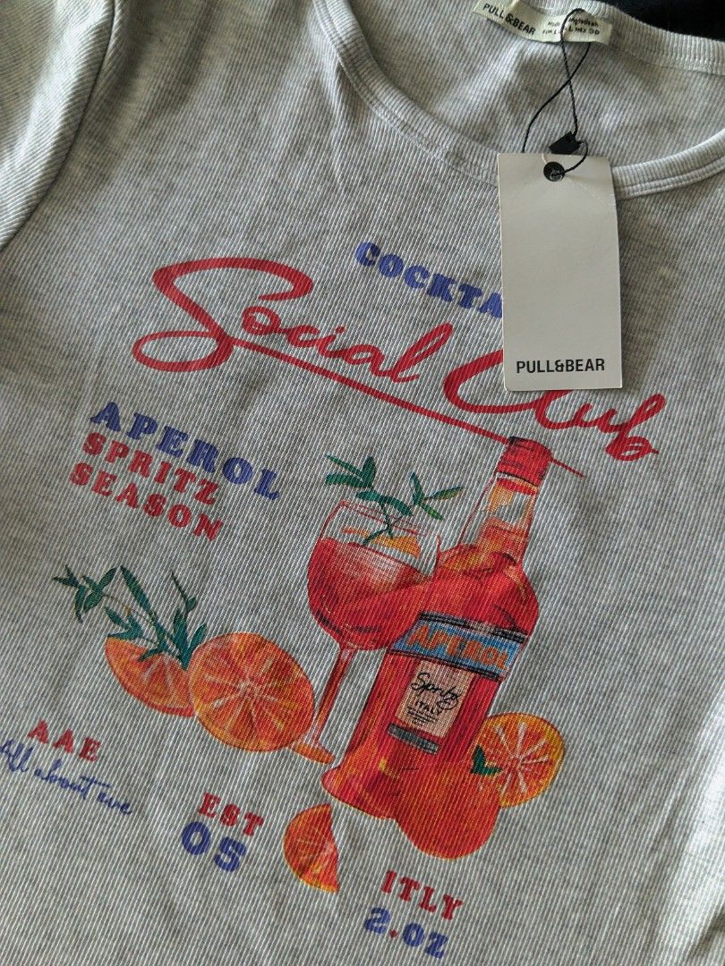Pull & Bear Aperol Spritz Graphic Ribbed T-Shirt (Category: Shirts) located in Kananga. Submitted by aurylien.clo (Listing ID 1467612513, Image 2). Description: - Brand: Pull & Bear - Condition: Brand new with original store tag attached - Color: Light grey - Material: Comfortable ribbed cotton - Design: Cute Aperol Spritz graphic with bottle, cocktail glass, and orange slices - Details: Short sleeves, scoop neckline, red and blue lettering.