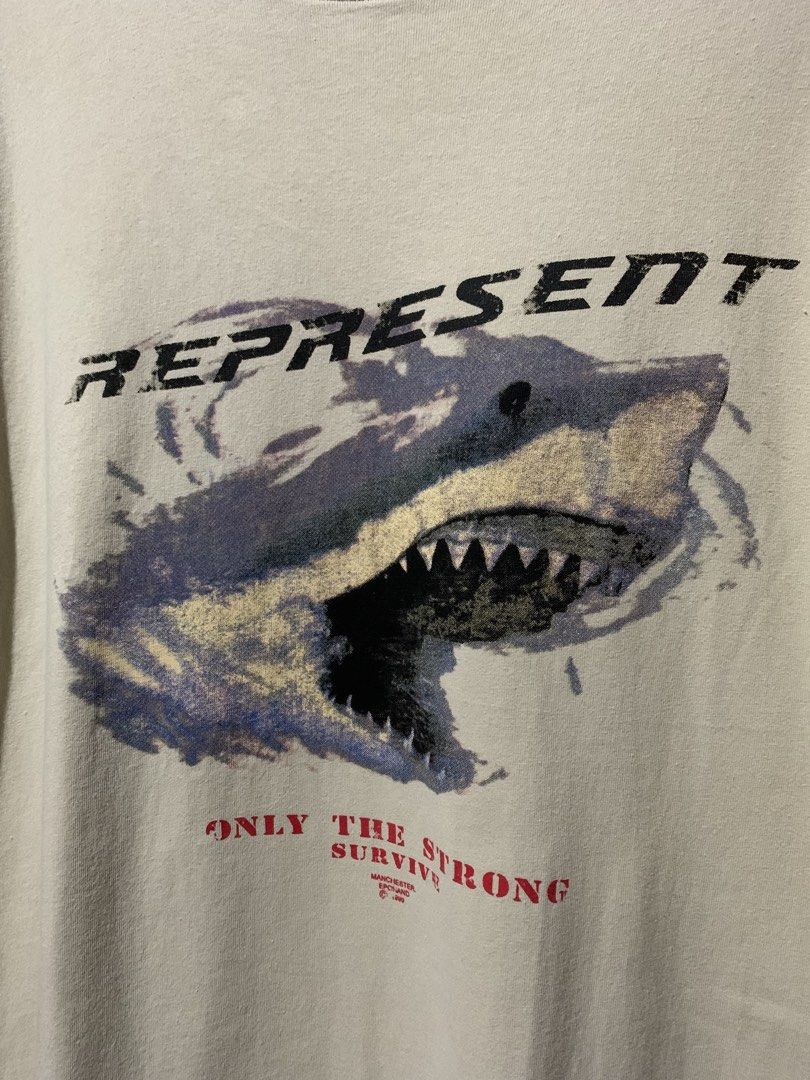 Represent Shark Only The Strong Survive Tee (Category: Tshirts & Polo Shirts ) located in Tayug. Submitted by cvcthrift (Listing ID 1467805723, Image 2). Description: 24/30 NO ISSUE EXCELLENT CR-9.5/10 - Brand: Represent - Color: Cream - Size: Medium - Material: 100% cotton - Design: Front shark graphic with 'Only The Strong Survive' text in red - Details: Distressed collar and signature metal Represent tag near the hem - Condition: Includes inne