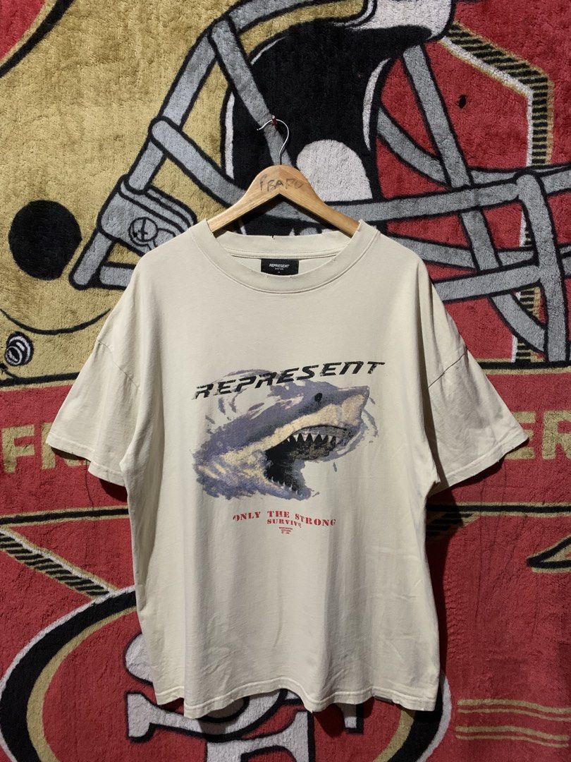 Represent Shark Only The Strong Survive Tee (Category: Tshirts & Polo Shirts ) located in Tayug. Submitted by cvcthrift (Listing ID 1467805723, Image 1). Description: 24/30 NO ISSUE EXCELLENT CR-9.5/10 - Brand: Represent - Color: Cream - Size: Medium - Material: 100% cotton - Design: Front shark graphic with 'Only The Strong Survive' text in red - Details: Distressed collar and signature metal Represent tag near the hem - Condition: Includes inne