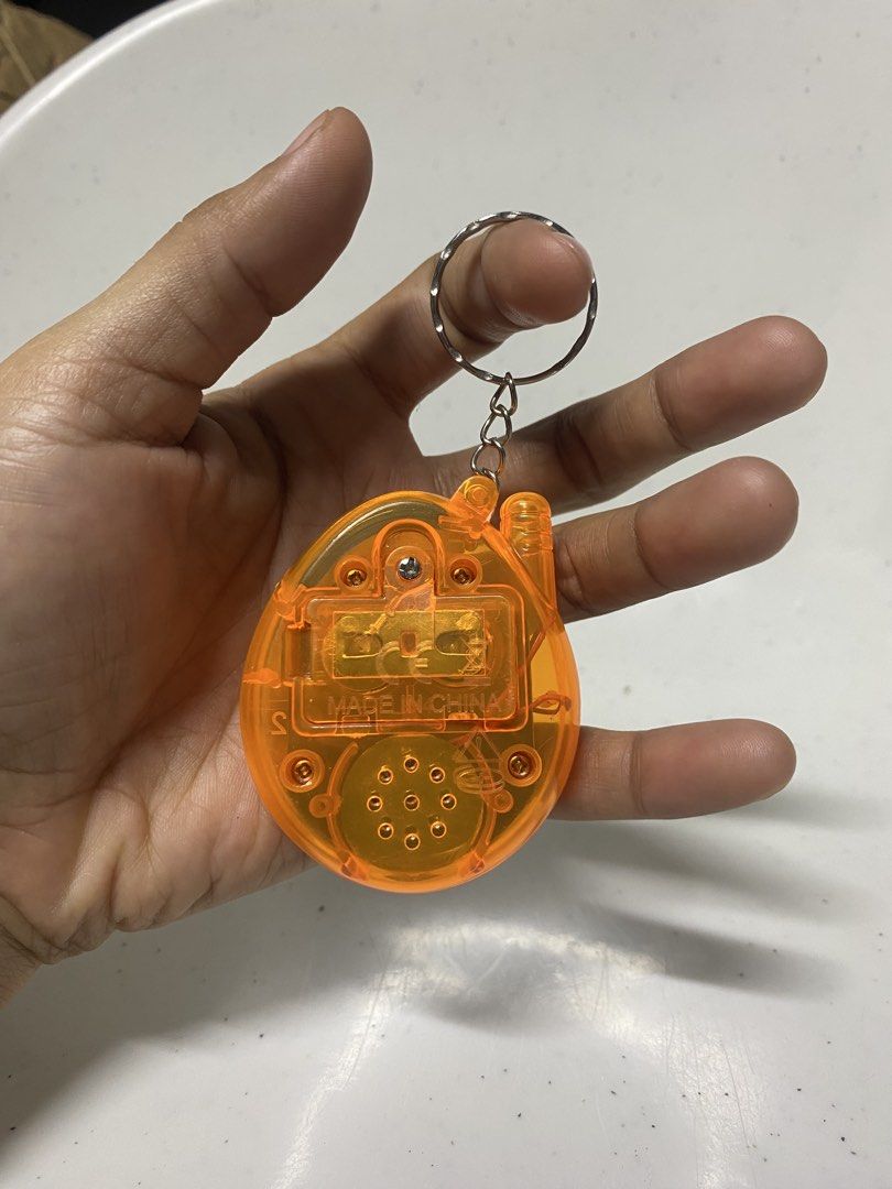 Transparent Orange Electronic Virtual Pet Game Keychain (Category: Interactive Toys) located in Pasacao. Submitted by wengpullar37205 (Listing ID 1467832863, Image 2). Description: - Orange translucent virtual pet keychain - Classic egg shape with blue buttons and antenna - Screen displays "PET GAME" in red font - Back features battery compartment with screws and speaker grille - Made in China -lagyan nio nlng po ng batt.wla aq bilog na batt.kaya