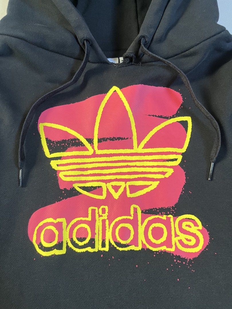 Adidas Black Graphic Trefoil Pullover Hoodie (Category: Hoodies) located in Candaba. Submitted by tristanpelayoo (Listing ID 1467857764, Image 2). Description: - Brand: Adidas - Style: Pullover hoodie with kangaroo pocket and adjustable drawstring hood - Color: Black with vibrant pink and yellow graffiti-style trefoil logo and sleeve graphics - Size: Asia M, AU/NZ M, US M, Korea XL - Condition: Pre-loved with noticeable fraying and a small tear a