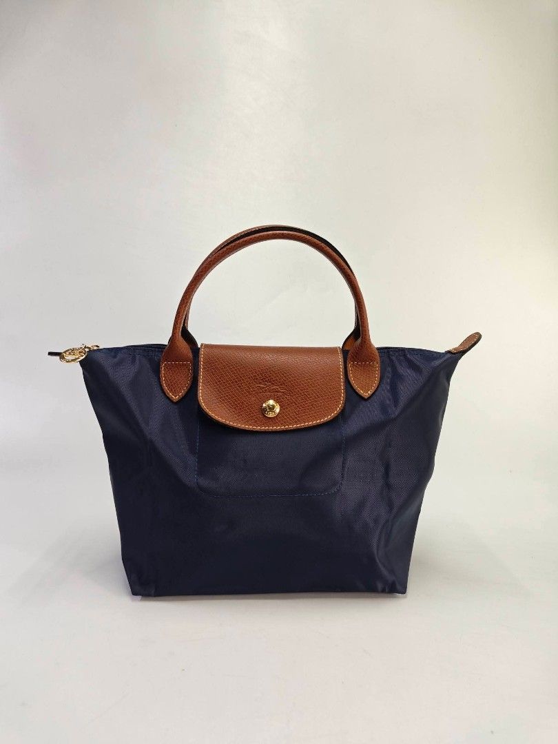 Authentic Longchamp Le Pliage Classic Small Short Handle Handbag - Navy (Category: Purses & Pouches) located in Quezon City. Submitted by hollowwillow (Listing ID 1468014219, Image 1). Description: - Brand/Model: Longchamp Handbag - Color: Classic Navy Blue with Brown Leather Flap and Handles - Hardware: Gold-tone - Closure: Zipper top with snap button flap - Interior: Dark lining with a convenient slip pocket - Style: Short handle design.
