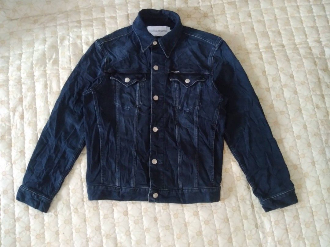 Calvin Klein Jeans Dark Blue Denim Jacket (Category: Coats, Jackets and Outerwear) located in Zamboanga. Submitted by nurbya898048 (Listing ID 1468057833, Image 1). Description: Dark blue denim jacket from Calvin Klein Jeans. Size S tag attached near the collar. Metal button closures down the front and on chest pocket flaps. Small branded tab on the left chest pocket. Classic collar style with contrast stitching throughout. Flat dimensions; Tag s