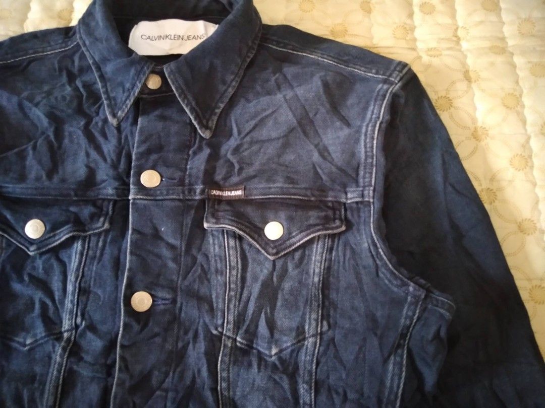 Calvin Klein Jeans Dark Blue Denim Jacket (Category: Coats, Jackets and Outerwear) located in Zamboanga. Submitted by nurbya898048 (Listing ID 1468057833, Image 2). Description: Dark blue denim jacket from Calvin Klein Jeans. Size S tag attached near the collar. Metal button closures down the front and on chest pocket flaps. Small branded tab on the left chest pocket. Classic collar style with contrast stitching throughout. Flat dimensions; Tag s