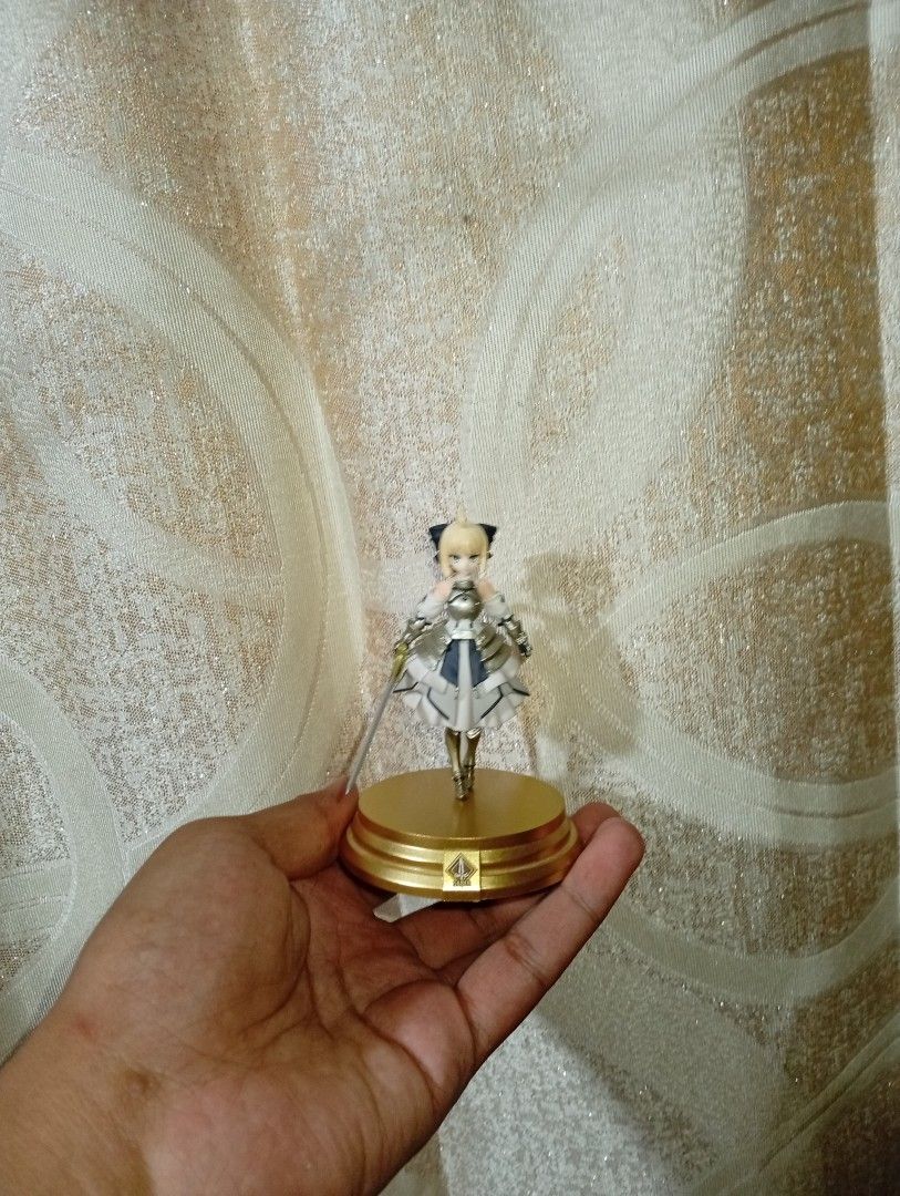 Fate/Grand Order Duel Saber Lily (Category: Figurines & Statues) located in Manila. Submitted by artisan027 (Listing ID 1467961147, Image 1). Description: - Character: Saber (Fate/Grand Order) - Condition: Complete with all original parts - Included Accessories: Golden circular display base, clear support rod, and detailed sword - Details: Blonde hair with black ribbons, detailed white and silver armor dress with ruffled skirt - Authenticity: Fea