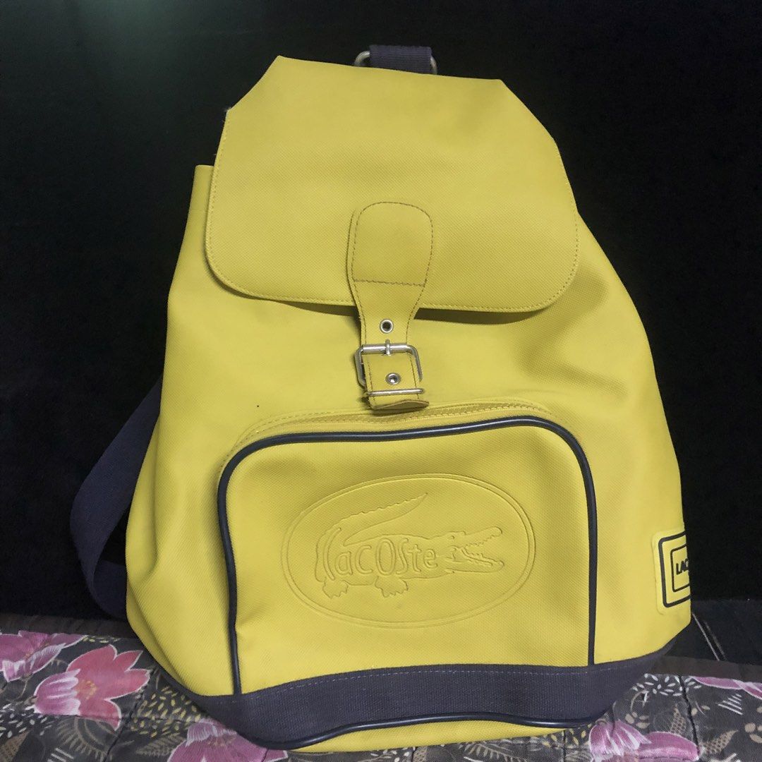 Lacoste Yellow Canvas Backpack with Embossed Crocodile Logo (Category: Backpacks) located in Malabon City. Submitted by jillmolo (Listing ID 1467960984, Image 3). Description: TAKE ALL Bright yellow Lacoste backpack made from sturdy canvas material. Features an embossed crocodile logo inside an oval patch on the front pocket. Flap closure with adjustable metal buckle strap. Dark blue strap and bottom panel contrast nicely against the yellow fabri