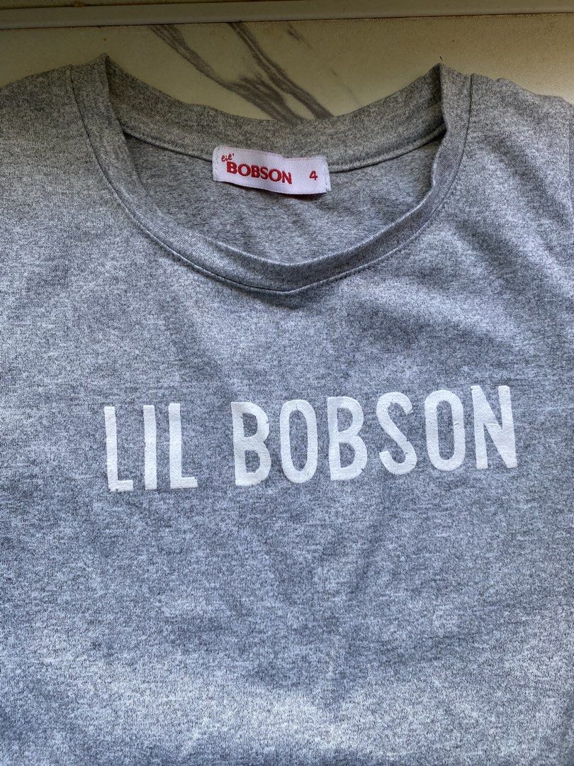 Lil Bobson grey t-shirt size 4 (Category: Shirts) located in Manila. Submitted by piinnnnkk (Listing ID 1467918089, Image 2). Description: - Brand: Lil Bobson - Size: 4 - Color: Grey - Material: Soft knit fabric blend - Design: White graphic text on chest with a cute tie detail at the bottom hem - Details: Crew neckline, standard hem, and signature red brand tags on the collar and lower side seam - Condition: Excellent, well-maintained.