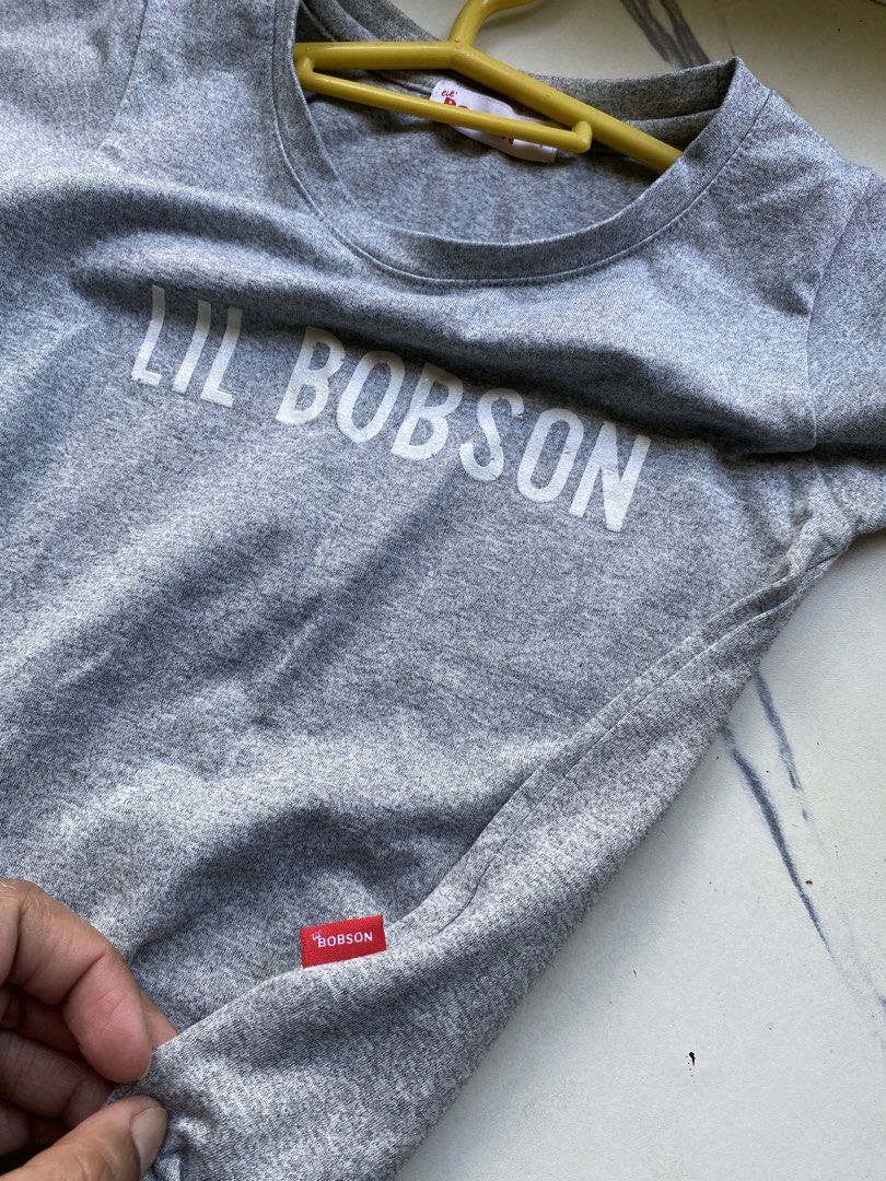 Lil Bobson grey t-shirt size 4 (Category: Shirts) located in Manila. Submitted by piinnnnkk (Listing ID 1467918089, Image 3). Description: - Brand: Lil Bobson - Size: 4 - Color: Grey - Material: Soft knit fabric blend - Design: White graphic text on chest with a cute tie detail at the bottom hem - Details: Crew neckline, standard hem, and signature red brand tags on the collar and lower side seam - Condition: Excellent, well-maintained.